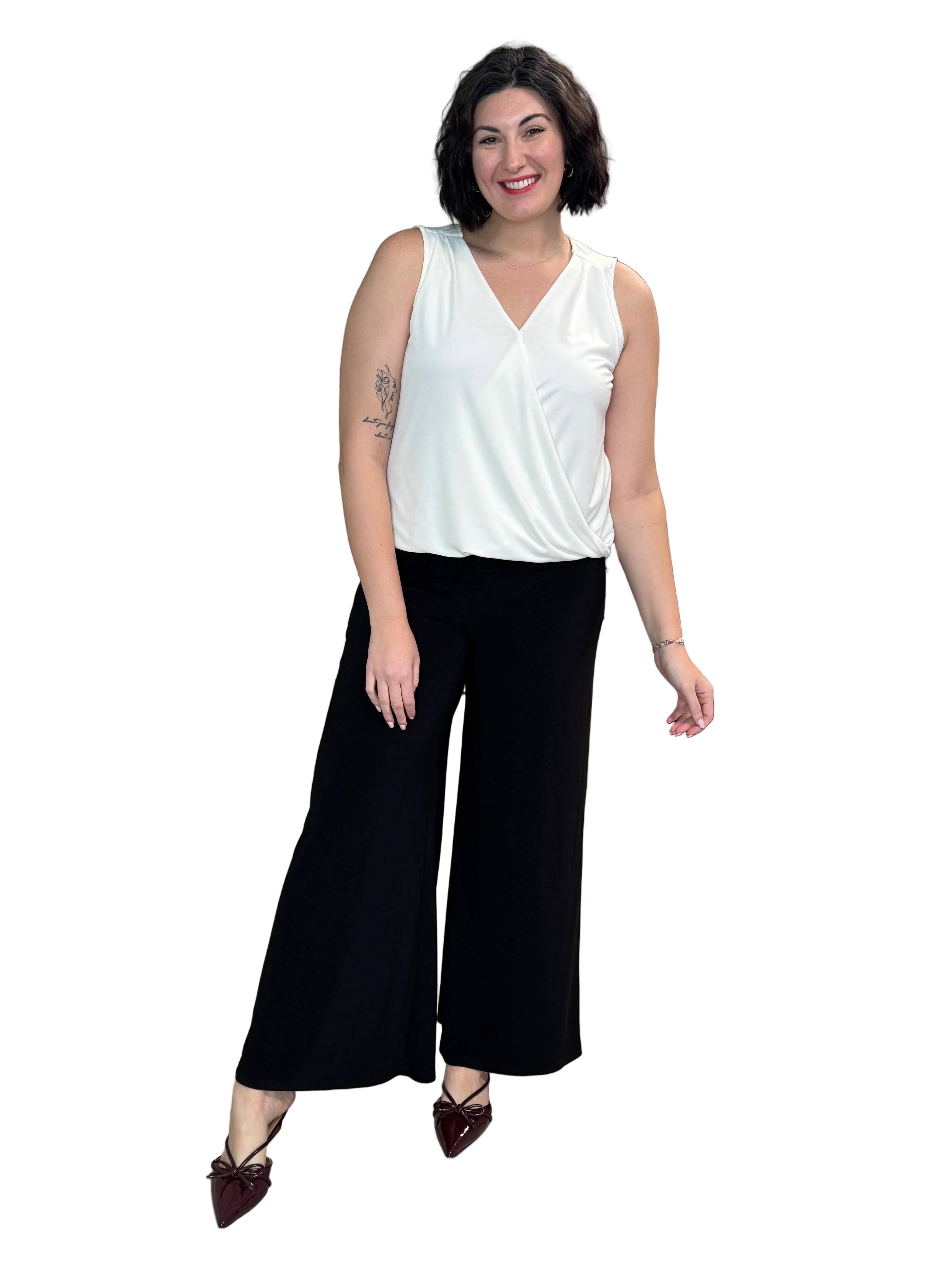 Woman in a white top and black pants standing in a room with a couch and decorative items.